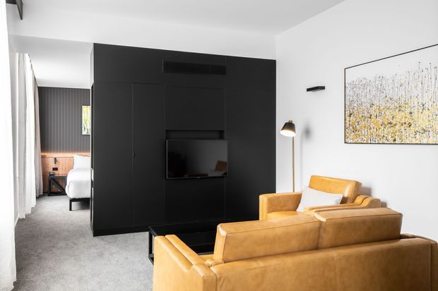 Contemporary living room with a warm tan leather sectional sofa and armchair facing a large black built-in media wall. The room has white walls and ceiling, grey carpet, a slim floor lamp, abstract artwork, and full-height curtains. A bedroom is visible through the opening at the rear, with dark vertical wall panelling and a timber-toned bedside area.