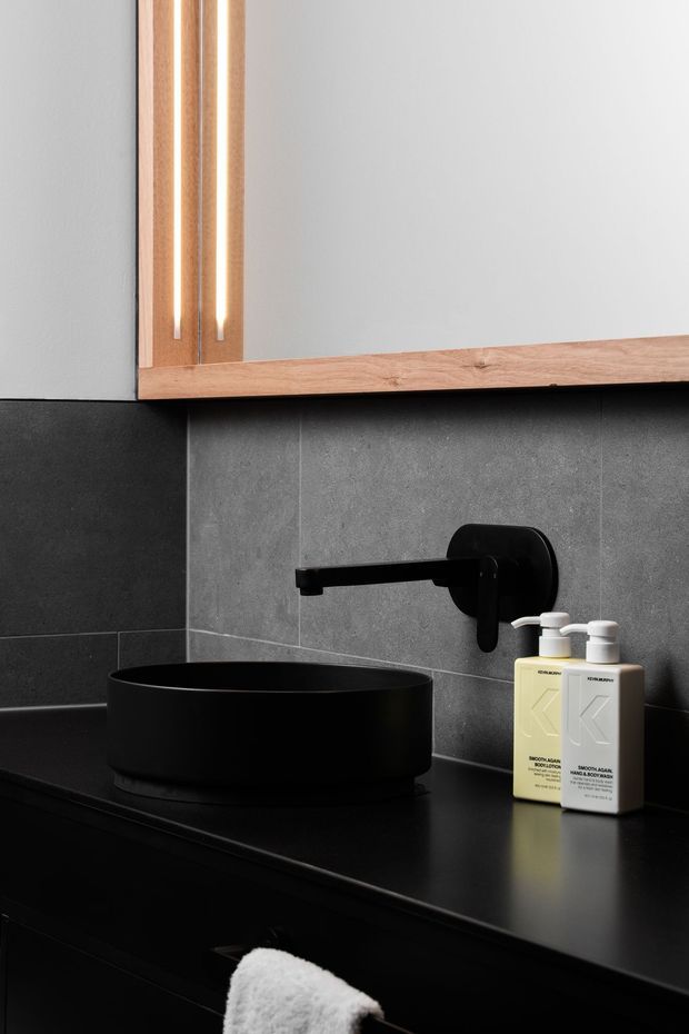 Close-up of a refined dark bathroom vanity with a matte black vessel basin, wall-mounted black tapware, dark stone or porcelain tiled splashback, and black benchtop cabinetry. A timber-framed mirrored cabinet with integrated vertical lighting is mounted above the basin, with neutral soap dispensers beside it. The composition is moody, minimalist, and high-contrast.
