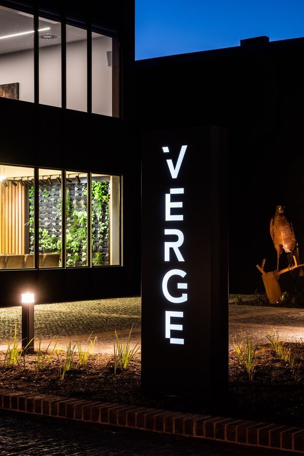 A dusk exterior view of a contemporary commercial building entrance. A tall black illuminated sign spells “VERGE” in white lettering, set within landscaped garden beds edged with dark brick. Behind it, floor-to-ceiling glazing reveals a brightly lit interior with a green living wall, timber screening, seating, and linear ceiling lighting. The dark facade and dramatic architectural lighting create a bold nighttime presentation.