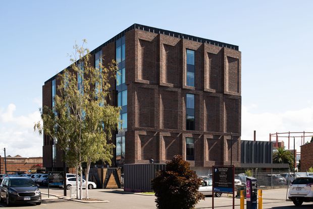 Exterior view of a contemporary multi-storey brick building with a strongly articulated geometric facade. Deep vertical brick reveals frame tall windows across the front, while a glazed side elevation provides contrast. The building fronts a paved urban street with mature trees, signage, fencing, and neighbouring commercial or industrial structures.