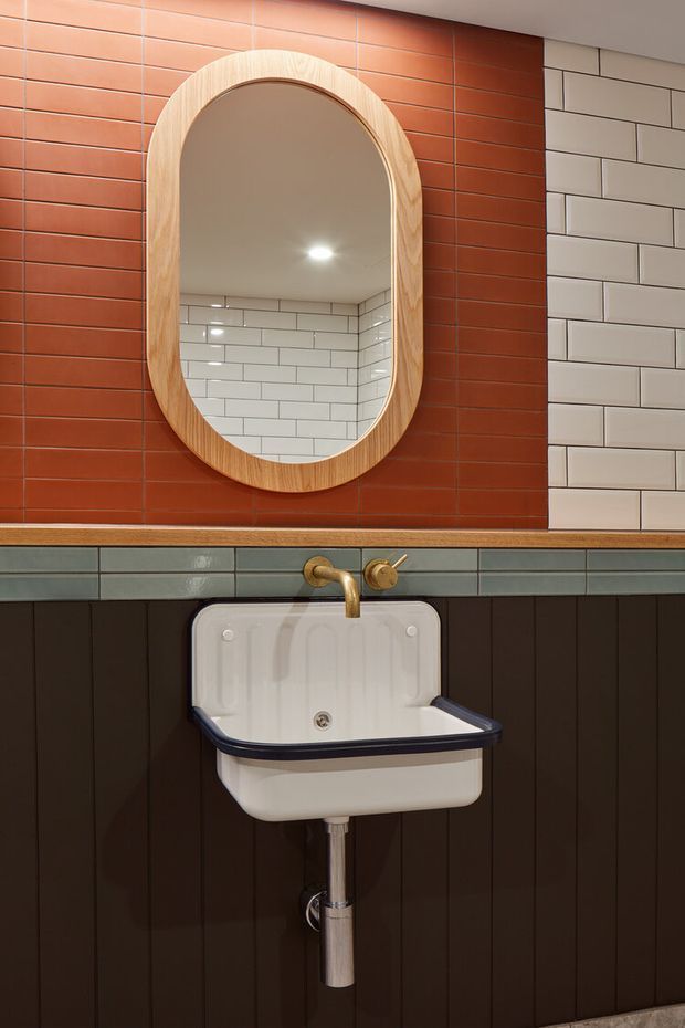 A compact, characterful bathroom or powder room centred on a wall-mounted white ceramic basin with dark trim and exposed chrome plumbing. The walls combine dark brown vertical wall panelling, a horizontal band of muted blue-grey glazed tiles, terracotta-red rectangular wall tiles, and white subway tiles. A rounded-arch timber-framed mirror sits above the basin, with brass/gold-toned wall-mounted tapware and warm recessed lighting. The composition has a refined retro feel with strong material and colour contrast.