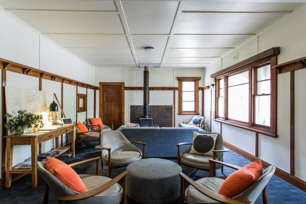 A bright, wood-trimmed living room with a central fireplace, blue sofa, mid-century lounge chairs, round ottoman, and a compact desk workspace. Large timber-framed windows line the right wall, while dark timber beams and trim contrast with the white paneled walls and ceiling.