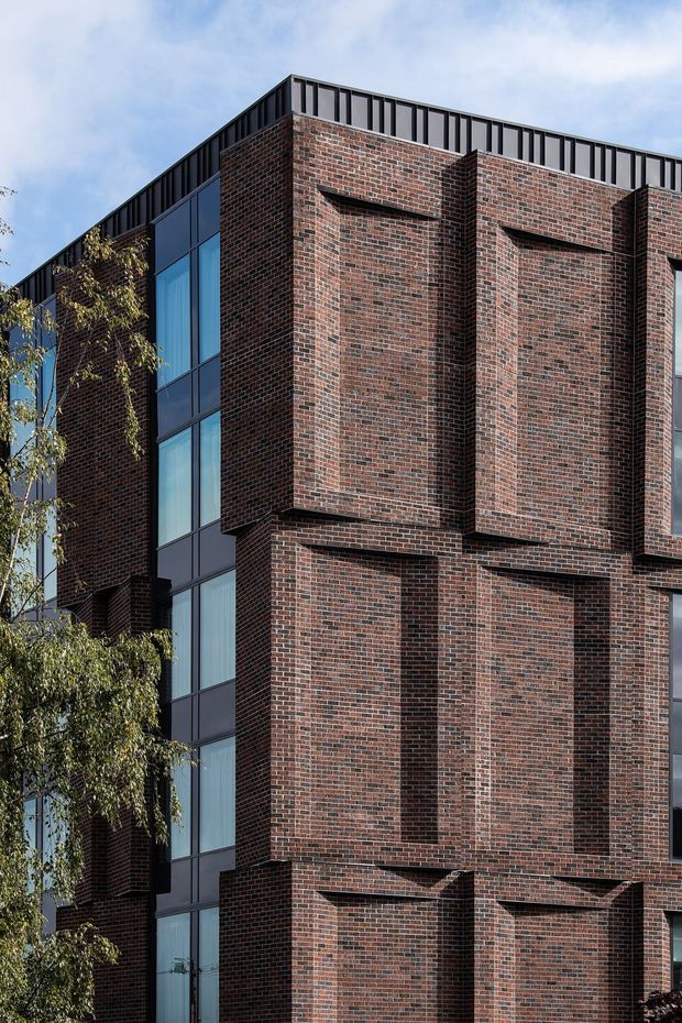Close-up exterior view of a multi-storey contemporary building with dark red-brown brick facade, strong projecting brick bays and recessed openings, tall dark-framed glazing, and a dark parapet or roof-edge screen. Trees partially frame the lower left, with sky in the background.