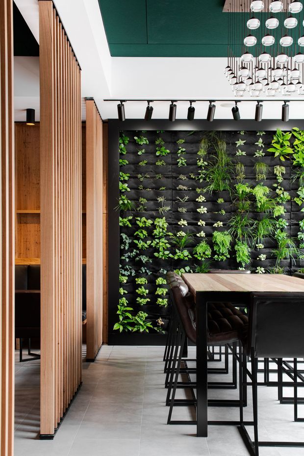 A contemporary commercial dining or hospitality interior with a long communal timber-topped table, black metal bar stools, and a dramatic living green wall. Vertical timber slatted screens divide the space, while a dark green ceiling, black track lighting, and a clustered pendant light installation create a refined, modern atmosphere. Large-format grey floor tiles and black built-in elements complete the scheme.