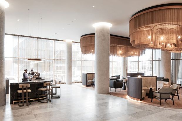 A spacious, refined commercial hospitality interior with a bar and dining area on the left, lounge seating on the right, expansive floor-to-ceiling glazing, round structural columns, and distinctive oversized circular pendant chandeliers with timber-toned slatted shades. The palette is light and neutral with warm wood and soft furnishings.