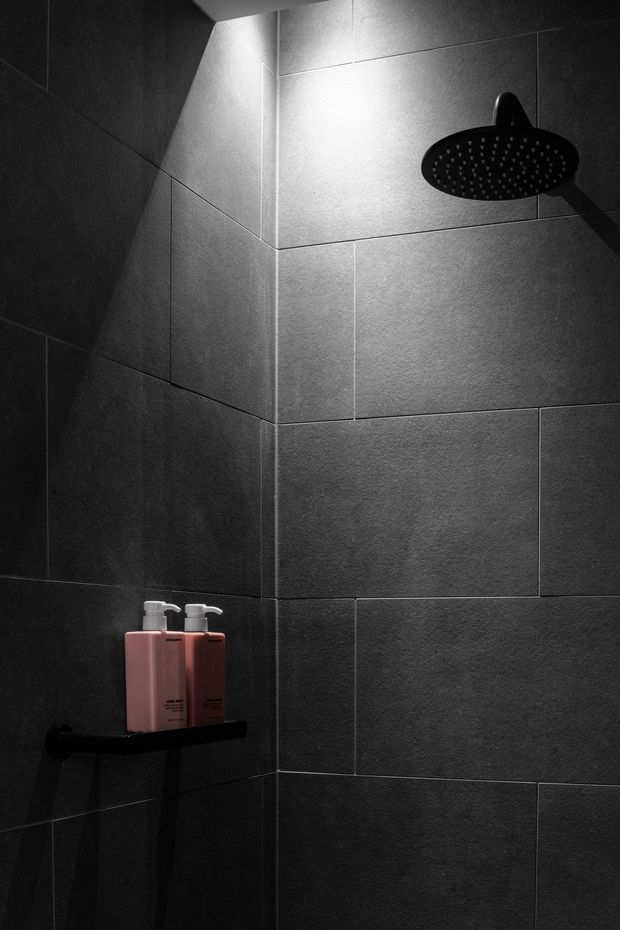 A close-up view of a dark, moody shower enclosure. Large-format charcoal grey textured wall tiles cover the walls, with a matte black round rainfall showerhead and black wall-mounted shelf. Two pink and dark red toiletry bottles provide a small colour accent. Dramatic directional lighting emphasizes the tile texture and corner junction.