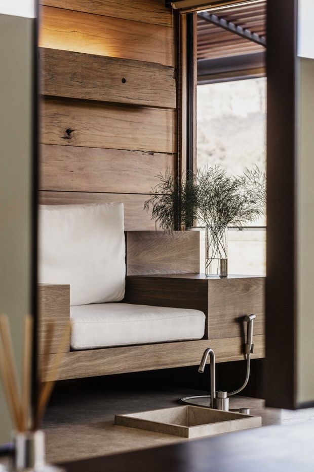 A close-up interior view of a compact, highly integrated kitchenette or wet-bar area beside a built-in upholstered bench. The space features warm, reclaimed-looking horizontal timber wall panelling and cabinetry, a dark wood counter, a small rectangular stone or concrete sink with a contemporary metal gooseneck tap and separate pull-out spray, and a narrow window with an exterior pergola visible beyond. A glass vase with delicate branches sits on the counter, adding a natural accent.