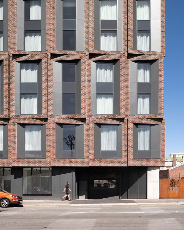 Street-level photograph of a contemporary multi-storey urban building facade. The elevation is composed of repeated projecting brick window bays with dark grey metal-framed glazing and light curtains, above a darker ground-floor frontage with a glazed entry, pedestrian door, and garage opening. Adjacent low-rise brick buildings and a clear blue sky are visible.