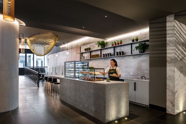 A sophisticated contemporary café interior with a long stone service counter, glass pastry display cases, open shelving with plants and crockery, and a barista station. The space features dark ceilings, textured concrete-like walls, tiled flooring, integrated linear lighting, pendant lights, and a stair or ramp leading toward bright glazing at the rear.