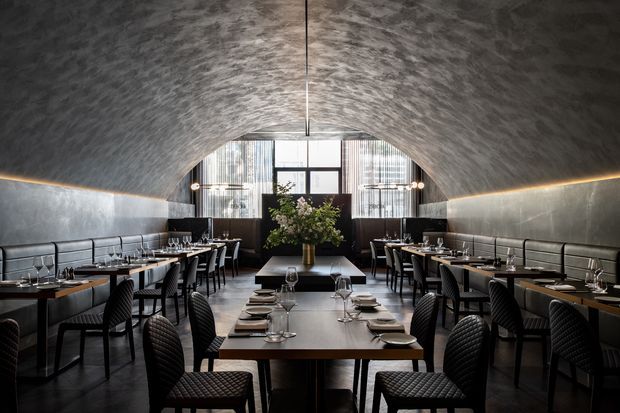 A sophisticated modern restaurant dining room with rows of neatly arranged dark tables and quilted upholstered dining chairs. Continuous banquette seating lines both walls beneath a dramatic barrel-vaulted, textured ceiling. Linear concealed lighting traces the wall and ceiling edges, while a large floral arrangement anchors the rear of the room in front of tall glazed windows. The palette is predominantly charcoal, black, grey, and dark timber with warm, atmospheric lighting.