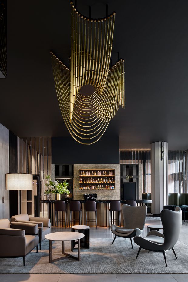 A sophisticated dark-toned hospitality lounge featuring a dramatic sculptural gold pendant chandelier, a polished bar with illuminated bottle shelving, high stools, and a seating area with upholstered armchairs and round coffee tables. Floor-to-ceiling windows, vertical timber detailing, stone-look wall surfaces, and layered ambient lighting create a refined, luxurious atmosphere.