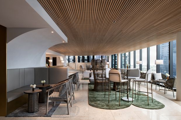 A sophisticated contemporary hospitality-style interior combining a lounge, bar, and dining area. The space features a sweeping ribbed timber ceiling, curved white plaster forms, floor-to-ceiling glazing, dark built-in banquette seating, sculptural tables, upholstered bar stools, and a mix of dining and lounge furniture arranged over green area rugs.