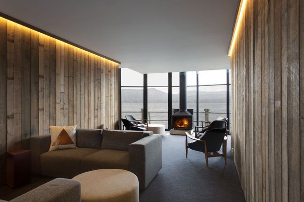 A contemporary living room with full-height glazing overlooking a calm mountainous or lakeside landscape. The room features dark timber wall panelling on both sides, warm concealed linear lighting, a large neutral sectional sofa, sculptural upholstered ottomans, mid-century-inspired lounge chairs, dark carpet, and a freestanding wood-burning fireplace centered in front of the windows.