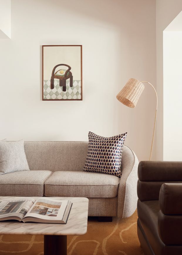 A warmly styled living room with a textured beige sofa, patterned accent cushion, dark brown leather armchair, marble-topped coffee table, woven floor lamp, framed artwork, and a soft geometric-patterned rug. The space has a calm, refined mid-century-inspired feel with warm neutral tones and carefully layered textures.