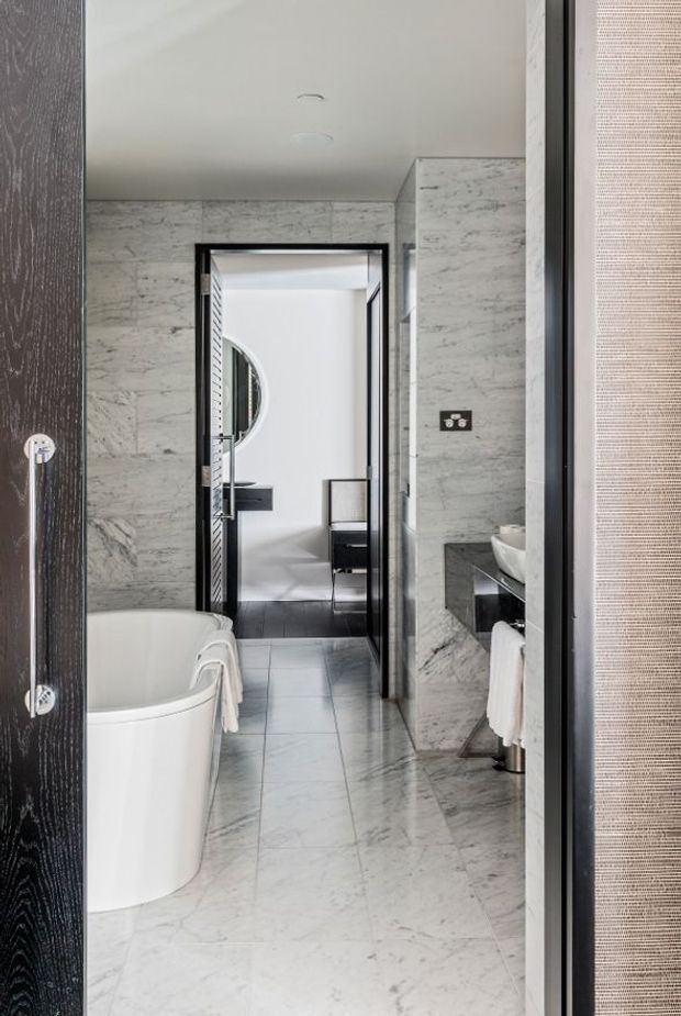 A refined contemporary bathroom clad in veined light-grey marble, featuring a freestanding white bathtub, marble vanity with a white basin, black-framed glazing and doors, and a view through to an adjacent bedroom or dressing area. The space has a cool, monochromatic palette with strong black accents and polished detailing.