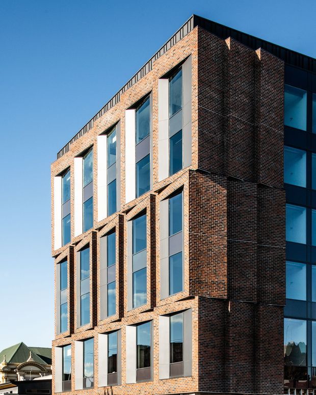 Exterior architectural photograph of a multi-storey urban building with a strong red-brown brick facade, vertically arranged floor-to-ceiling windows, projecting brick window bays, and dark metal detailing along the roofline and side elevation. A glassy dark-framed volume is visible at the right edge, with a neighbouring heritage-style roof partially visible in the background. Clear blue sky and crisp sunlight emphasize the building's geometric composition.