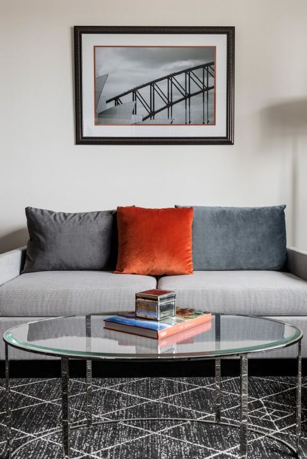 A refined living room vignette featuring a light grey upholstered sofa with charcoal and orange cushions, a large oval glass coffee table with a dark metal frame, and a geometric black-and-white rug. A framed architectural bridge photograph hangs above the sofa, with neutral walls and understated contemporary furnishings.