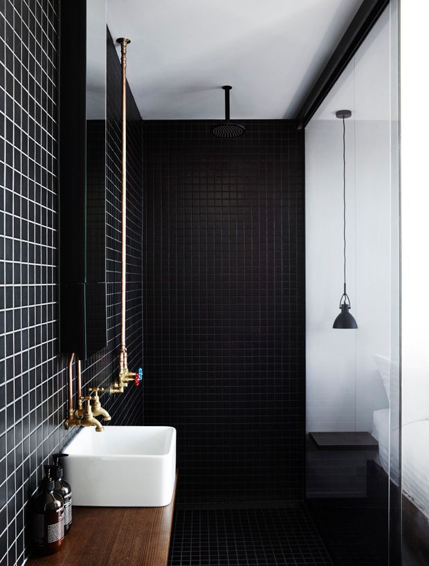 A dramatic, narrow contemporary bathroom with a walk-in shower clad in small matte black grid tiles. A white vessel basin sits on a warm timber vanity with exposed brass plumbing and black wall-mounted fittings. A black rain shower, black pendant light, glass partition, and a glimpse of a white bed beyond the glazing create a moody, hotel-like atmosphere with strong contrast between black tile, white fixtures, timber, and brass.