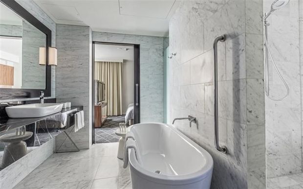 Luxurious marble-clad bathroom featuring a freestanding white bathtub, large wall mirror, double vessel vanity, wall-mounted sconces, and dark metal fixtures. A bedroom or dressing area is visible through the open doorway.