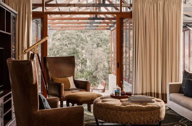 Warm, richly furnished living room with multiple brown leather lounge chairs, a large tufted ottoman, a pale sofa, dark built-in cabinetry, and full-height beige curtains. Timber-framed glazing and an open glass door reveal a leafy wooded setting, while exposed timber roof framing creates a strong indoor-outdoor connection and architectural focus.