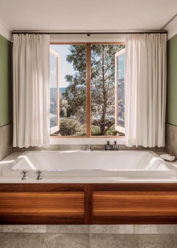 A refined bathroom centred on a large white built-in bathtub beneath an open timber-framed window. Full-height cream curtains frame the view, while muted green painted walls, stone-look wall and floor tiles, and rich dark timber vanity cabinetry create a warm, natural palette. Traditional chrome tapware and simple trim details add a classic character.