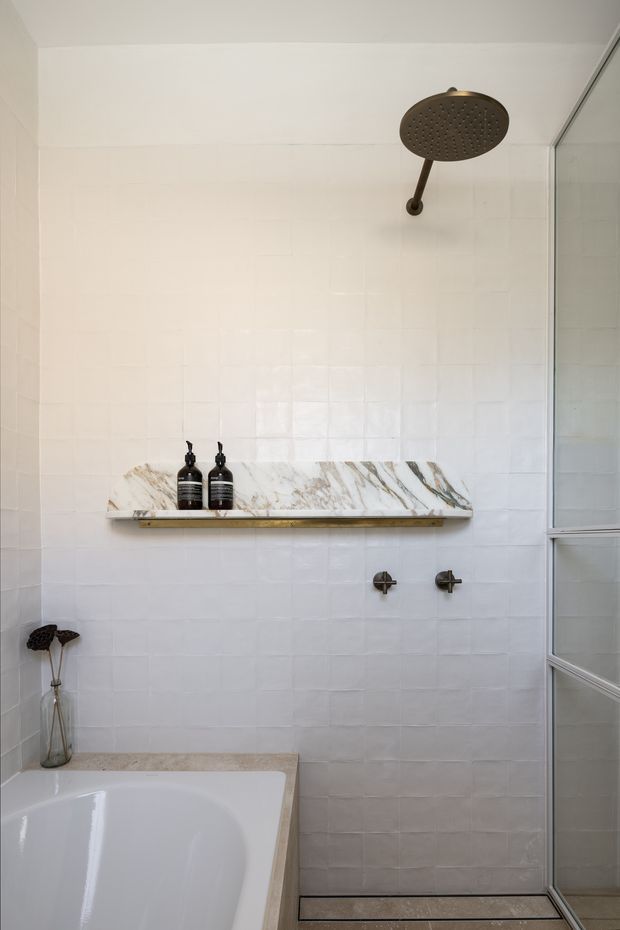 A bright contemporary bathroom featuring a white tiled walk-in shower with a large round dark metal rain showerhead, wall-mounted controls, and a marble-look stone shelf holding two dark bottles. A freestanding or built-in white bathtub with a pale stone surround occupies the lower left, while a framed glass shower screen is visible on the right. The palette is predominantly white and off-white with subtle grey and warm stone accents.
