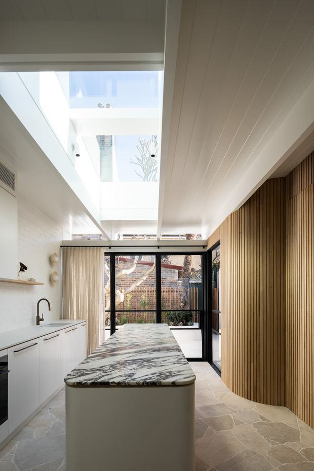 Bright, narrow contemporary kitchen with a large sculptural stone island, white cabinetry, black-framed glazed doors, and a dramatic central skylight. The kitchen combines a pale natural-stone floor with a white timber-lined ceiling, vertical timber slat feature wall, and a small courtyard visible beyond the glazing.