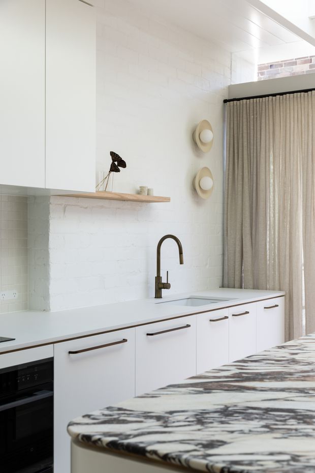 Bright contemporary kitchen with white cabinetry, a long pale benchtop, integrated appliances, a dark curved faucet, open timber shelving, and a dramatic patterned stone island benchtop in the foreground. White-painted brick walls, simple wall sconces, and full-height neutral curtains create a warm minimalist interior.