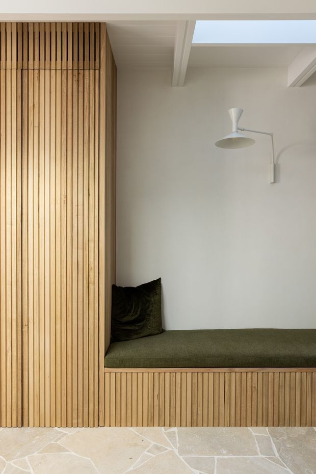 A minimalist built-in alcove seating nook with vertical light timber slat panelling and matching integrated bench cabinetry. The bench has an olive-green upholstered cushion and dark cushion, set against a warm off-white wall. A simple white wall-mounted reading lamp sits above, while exposed white ceiling beams and a skylight bring in natural light. The floor is finished in irregular pale stone slabs with light grout lines.