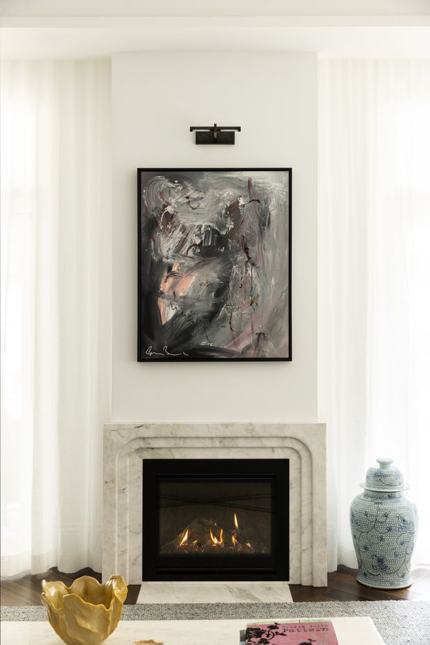 Bright, refined living room vignette centred on a contemporary fireplace with a carved pale stone surround, abstract artwork, and a black picture light. Sheer white curtains flank the fireplace, while a patterned ceramic urn and sculptural yellow vessel add decorative accents. A coffee table and rug are partially visible in the foreground.