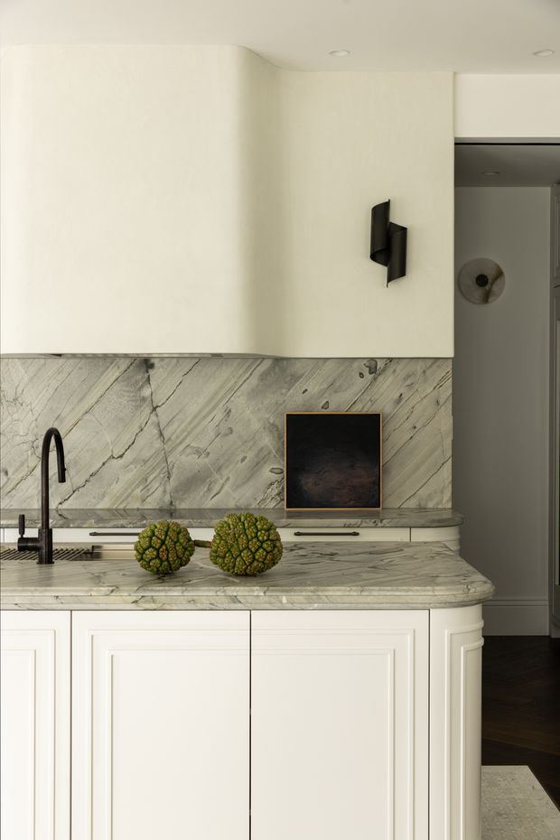 Close-up view of a refined kitchen featuring a pale veined stone splashback and benchtop, off-white shaker-style cabinetry, an integrated sink with a black gooseneck tap, dark hardware, and a small framed artwork. The kitchen opens toward a darker adjacent area at right, with warm timber flooring visible beyond.