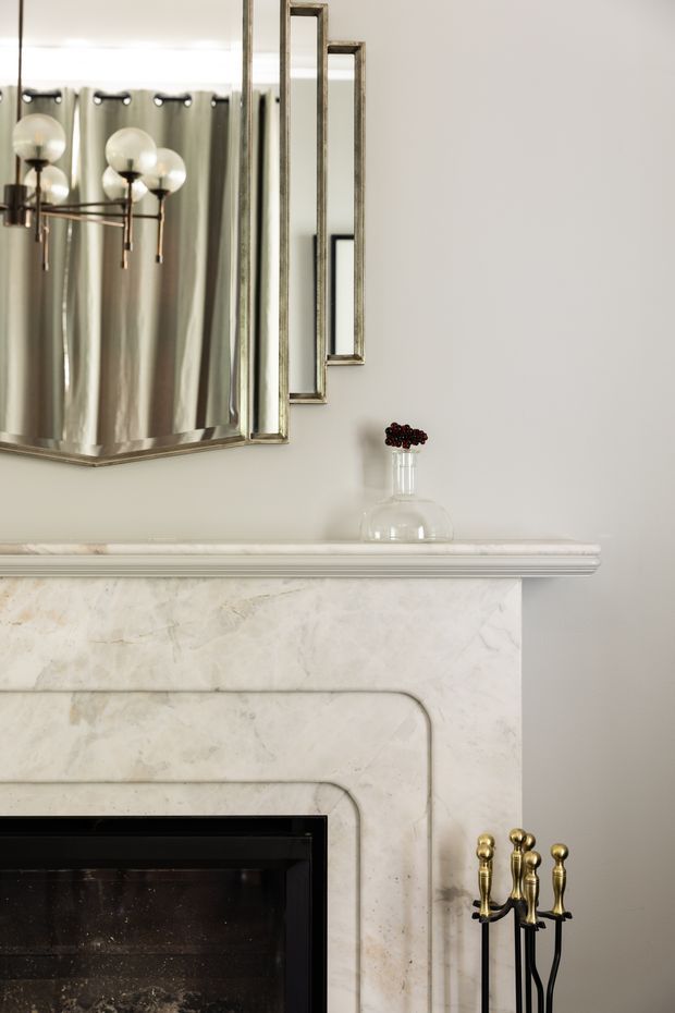 Close-up interior view of a refined traditional fireplace surround in pale veined marble, with layered curved detailing and a white moulded mantel shelf. Above it is a large aged metal-framed mirror reflecting a sculptural globe-bulb light fixture and draped curtains. Brass and black fireplace tools sit beside the dark firebox, while a small glass vase decorates the mantel.