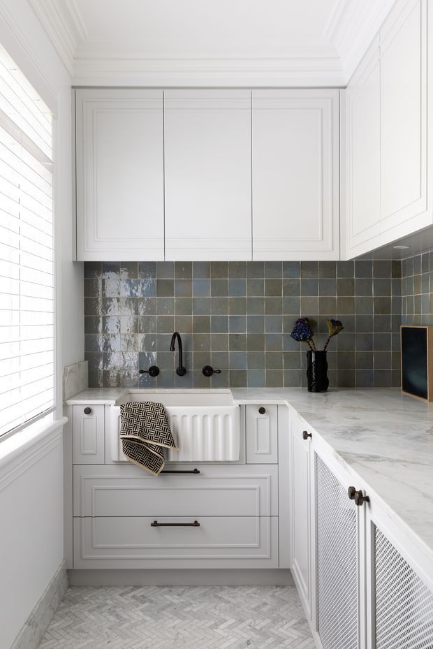 A bright, compact laundry room with white panelled cabinetry, a marble-look benchtop, integrated farmhouse-style sink, black tapware, and glossy grey-green square tiled splashback. The room features decorative moulding, white plantation shutters, and a light herringbone tile floor. A patterned towel and small vase provide contrasting accents.