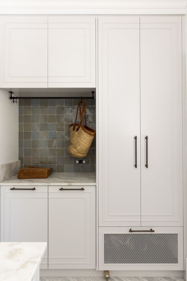 Close-up view of a refined white built-in kitchen or utility-room joinery wall. Shaker-style cabinetry with dark bar handles surrounds a recessed open storage nook featuring a grey-blue square tile backsplash, stone countertop, hooks, and a woven basket. A tall cabinet includes a decorative metal mesh ventilation panel near the base, with part of a pale marble-look benchtop visible in the foreground.