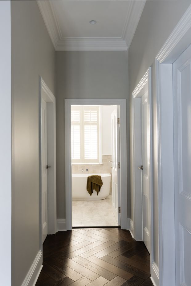 A refined residential hallway viewed toward a bright bathroom with a freestanding bathtub. The hallway has dark herringbone timber flooring, soft grey walls, white paneled doors and elaborate white crown moulding and trim. The bathroom beyond features pale flooring, shuttered windows, light wall finishes and a freestanding white tub. The image has a bright, calm, classic character.