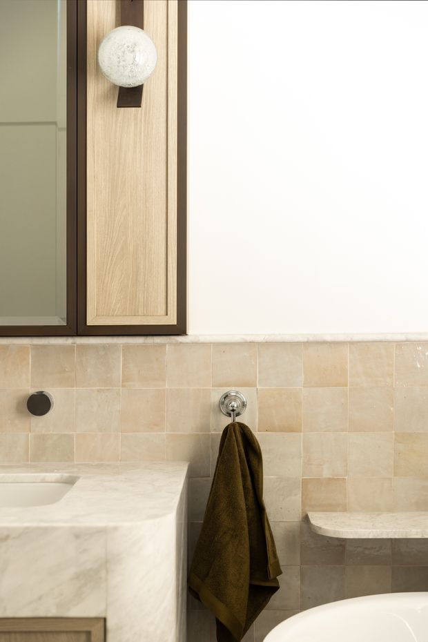 Close-up view of a refined bathroom vanity and bath area. The space features pale beige tiled walls, a substantial light marble vanity with an integrated basin, dark bronze or black tapware, a timber-framed mirrored cabinet with a spherical wall light, and a towel hanging beside the bathtub. The composition highlights layered natural materials and warm neutral finishes.