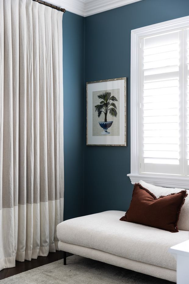 A refined sitting area with a cream upholstered chaise or sofa, dark timber flooring, a deep blue painted wall, white plantation shutters, full-height off-white curtains, and layered white crown moulding and window trim. A framed botanical artwork and rust-brown accent cushion add warmth and contrast.