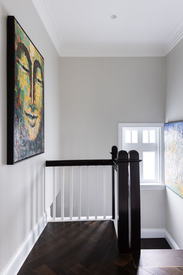 A bright residential stair landing with dark herringbone timber flooring, white painted walls and panelling, ornate white crown moulding, and a dark timber stair balustrade with white vertical spindles. A small multi-pane window provides natural light, while colourful abstract artwork hangs on the walls. The space has a refined traditional character with strong black-and-white contrast.