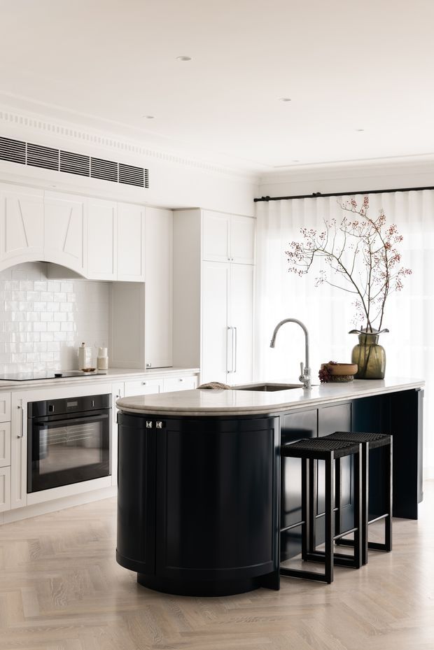 Bright, refined kitchen with white raised-panel cabinetry, a glossy white subway-tile splashback, pale timber flooring, and a prominent black curved island with a light stone benchtop. The island includes a sink and curved tap, with two black bar stools. Tall sheer curtains, decorative ceiling cornices, and a large vase with branches contribute to the elegant classic interior.