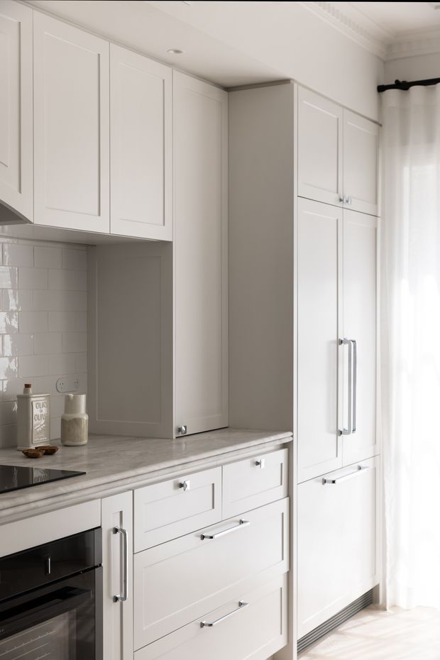 Bright, refined white kitchen cabinetry with shaker-style doors and drawers, integrated tall storage, stainless handles, a pale veined stone benchtop, and glossy white subway-tile splashback. Sheer curtains and daylight create an airy, softly traditional kitchen interior.