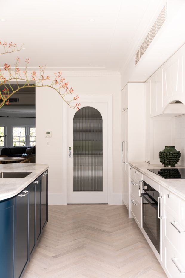 Bright, refined kitchen with white paneled cabinetry, dark blue island cabinetry, pale stone benchtops, and a light timber herringbone floor. The space features integrated appliances, a sculptural rangehood, decorative wall mouldings, a glazed arched door, and a view into an adjoining living area. A branch arrangement adds a subtle organic accent.