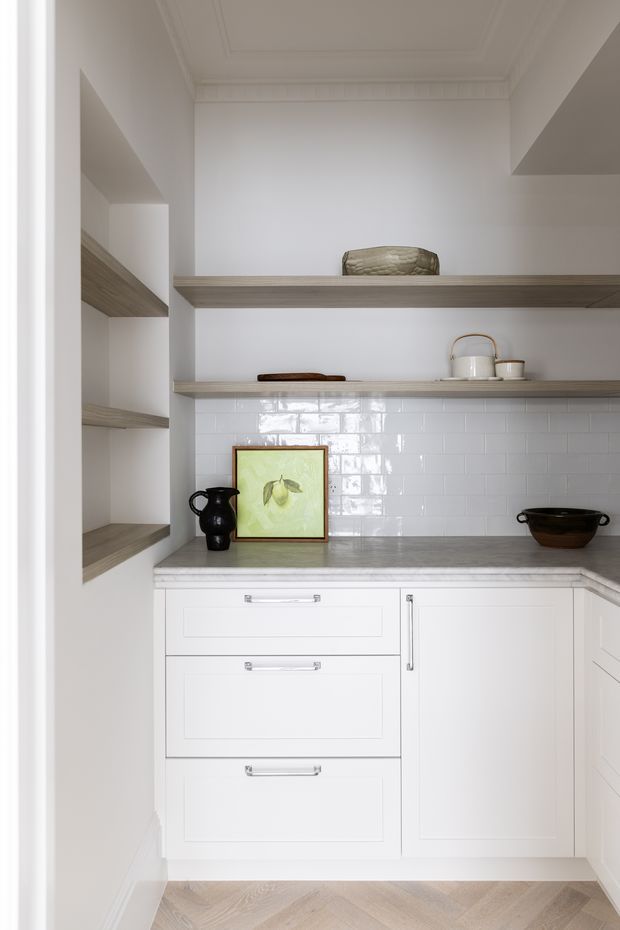 A compact, bright kitchen with white shaker-style cabinetry, pale timber open shelving, a grey stone-look benchtop, and glossy white subway-tile splashback. The space includes integrated drawers and cupboards, open display shelves, neutral decorative objects, and a light herringbone timber floor. Decorative ceiling mouldings add a refined traditional touch.