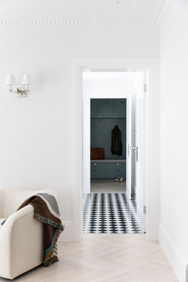 Bright white interior view from a living room or sitting area through a wide doorway into a dark teal built-in mudroom or entry hall. The foreground has pale herringbone timber flooring, white walls with detailed traditional mouldings, a wall sconce, and a cream upholstered armchair draped with a patterned throw. The adjoining space features full-height teal cabinetry, hooks, a bench, and a bold black-and-white geometric tile floor. The overall composition is crisp, refined, and high-contrast.
