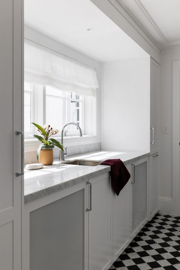 Bright, narrow white utility/laundry room with classic built-in cabinetry, a marble-look countertop, undermount sink and high-arched chrome tap beneath a window. White roller blinds, detailed cornice moulding, panelled doors, a vented cabinet panel and a black-and-white checkerboard floor create a refined traditional look. A potted plant and burgundy towel add small accents.