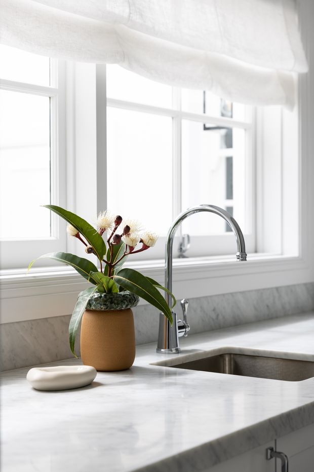 Bright, close-up view of a refined kitchen sink area. A polished marble countertop and backsplash surround a deep undermount sink with a tall chrome gooseneck tap and separate soap dispenser. Large white-framed windows with a sheer Roman blind provide abundant natural light, while a ceramic planter adds a small organic accent.