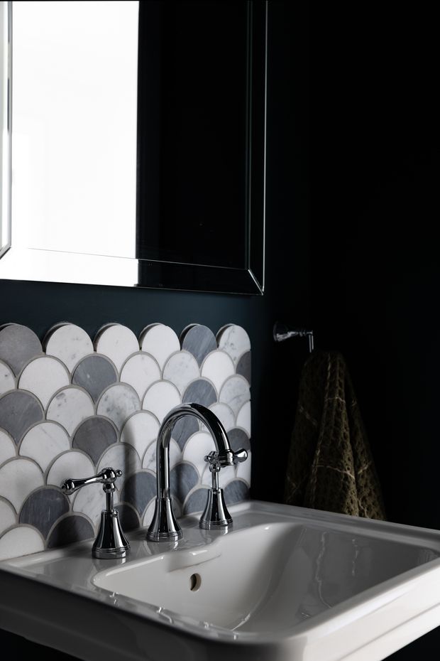 Close-up view of a sophisticated bathroom vanity featuring a glossy white ceramic basin with polished chrome three-piece tapware, set against a dark wall. The backsplash is clad in scallop-shaped mosaic tiles in white, pale grey, and charcoal tones, with a large black-framed mirror above and a dark textured hand towel hanging to the side. The space has a dramatic, high-contrast contemporary look.