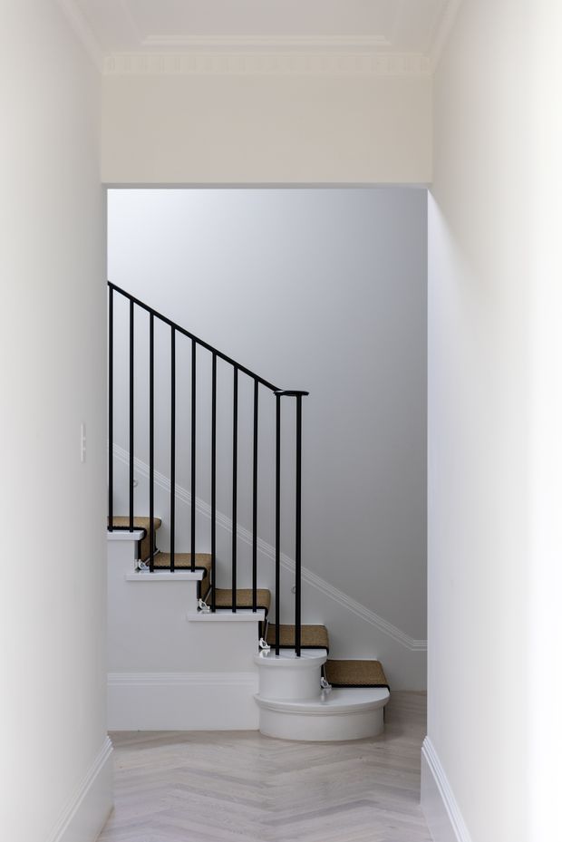 A bright, refined stairway viewed from a hallway, featuring a curved staircase with dark carpeted treads, white painted stepped sides and moulded detailing, and slim black metal balusters with a simple handrail. The surrounding walls and ornate ceiling cornice are painted off-white, while the adjoining wall is light grey with white trim. The floor is finished in pale herringbone timber.