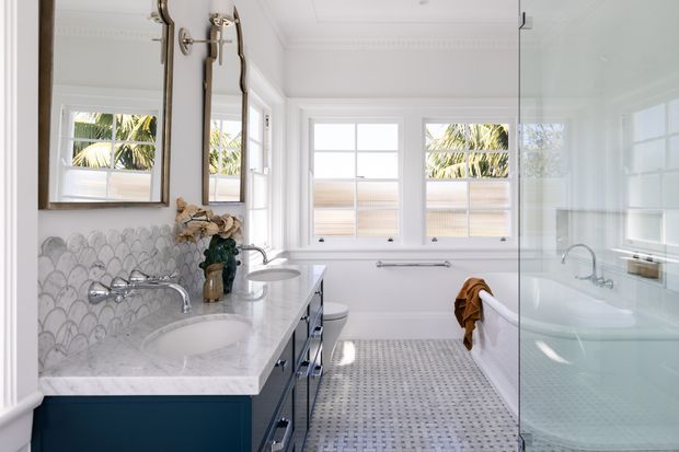 Bright, elegant bathroom with a double vanity, white marble-look benchtop, deep blue cabinetry, scalloped mosaic wall tile, freestanding bathtub, glass shower enclosure, and large white-framed windows. Brass-framed mirrors and polished chrome fixtures add refined detail, while palm foliage is visible outside.