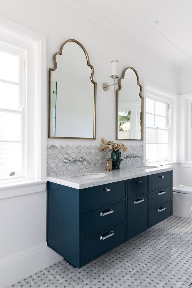 Bright, refined bathroom featuring a dark navy double vanity with marble countertop, two ornate arched mirrors, scalloped mosaic tile backsplash, polished chrome wall-mounted faucets, white trim, and a small-format patterned tile floor. Large white-framed windows provide abundant natural light, with a freestanding toilet visible at right.