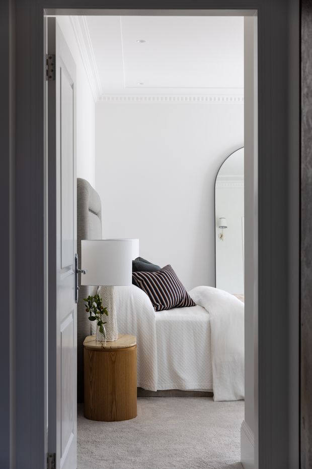 View through an open doorway into a bright, refined bedroom. The room has white walls and ceiling with decorative cornice moulding, pale carpet, a textured white bedspread, a grey upholstered bedhead, a striped dark accent cushion, and a rounded timber bedside table with a white lamp and small plant. A large arched mirror is visible against the far wall. The image is composed from the adjoining doorway, with the bedroom clearly dominating the view.